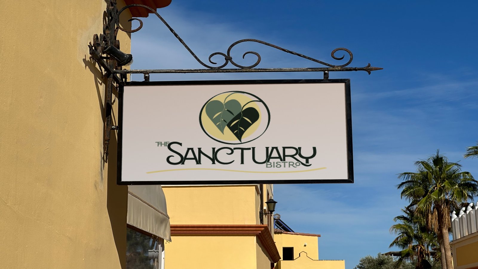 Wrought-iron hanging sign for The Sanctuary Bistro with the heart-leaf mark, mounted on an ochre wall against a blue sky with palm trees