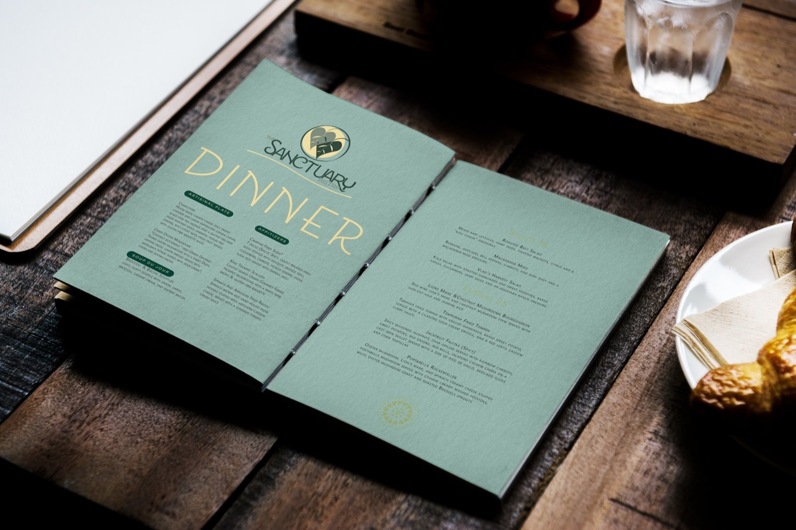 Sage green bifold dinner menu on a dark wood table set with a water glass and gold dish