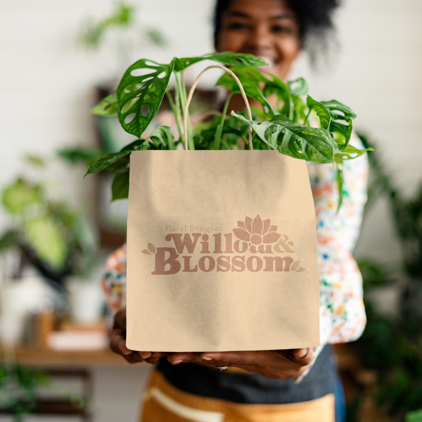 Smiling woman holding a kraft tote bag with the tonal Willow & Blossom lockup, planted with a trailing monstera